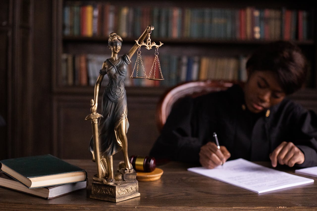 about-02 A focused judge writing on documents beside a Lady Justice statue in an office.